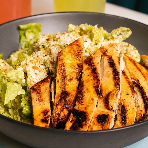 Grilled chicken Caesar salad in a dark bowl with drinks in the background.