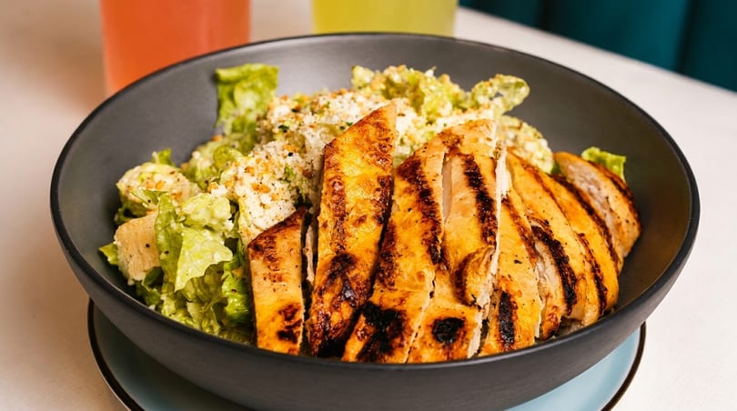 Grilled chicken Caesar salad in a dark bowl with drinks in the background.