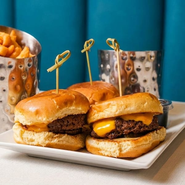 Three cheeseburger sliders with picks on a white plate, with a side of fries in a metal cup.