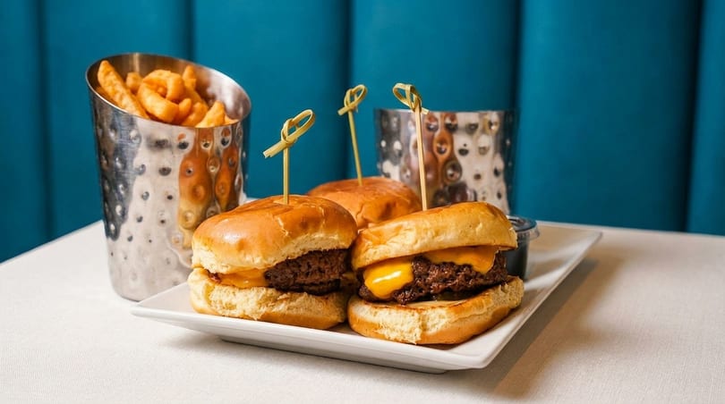 Three cheeseburger sliders with picks on a white plate, with a side of fries in a metal cup.