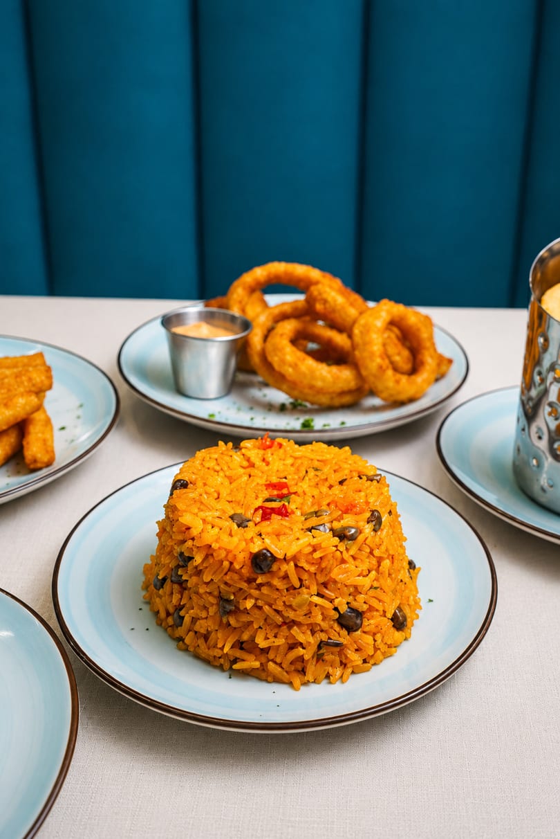 Plates of yellow rice with black beans, onion rings, and fried plantains on a white tablecloth.