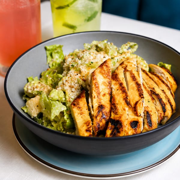 Grilled chicken Caesar salad in a dark bowl with two colorful drinks in the background.