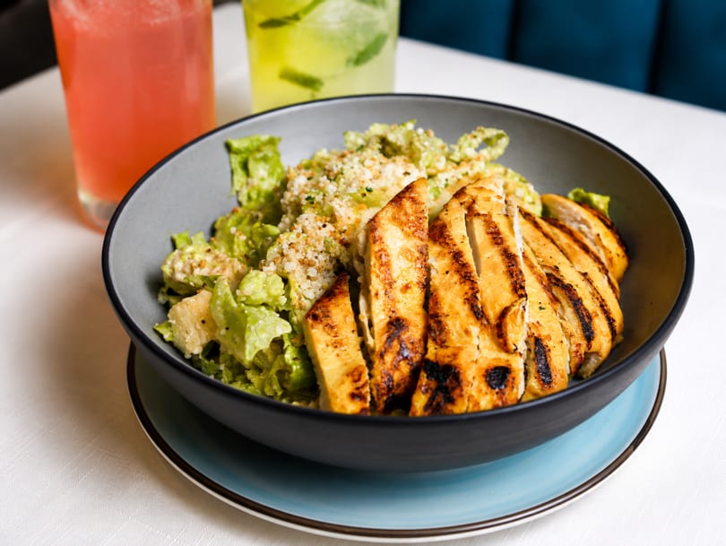 Grilled chicken Caesar salad in a dark bowl with two colorful drinks in the background.