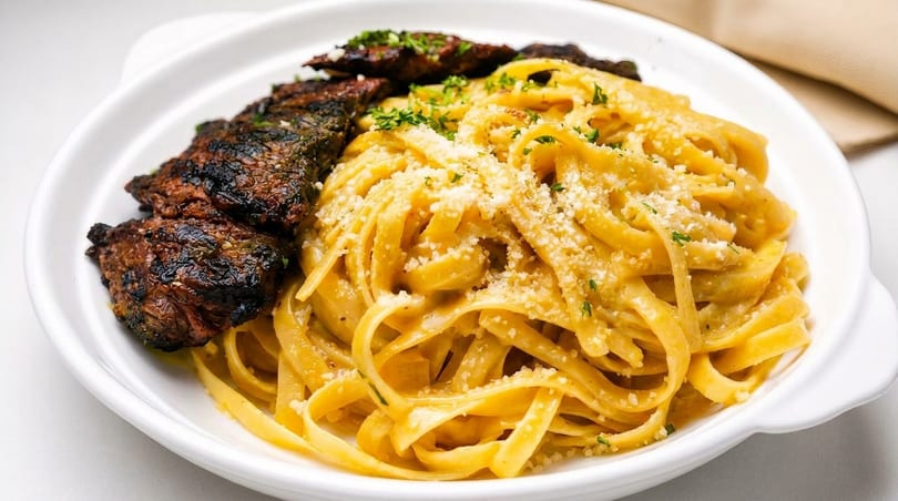 Grilled steak slices and creamy fettuccine pasta topped with parmesan cheese and parsley in a white bowl.
