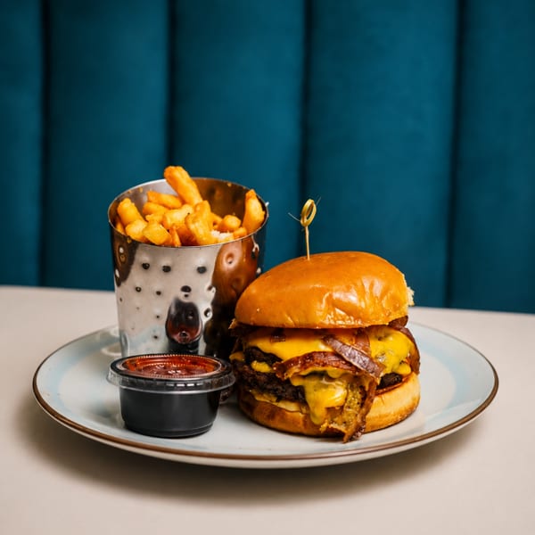 A double cheeseburger with bacon, caramelized onions, fries in a metal cup, and dipping sauce.