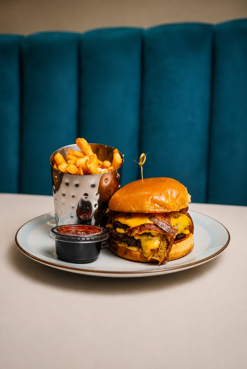A double cheeseburger with bacon, caramelized onions, fries in a metal cup, and dipping sauce.