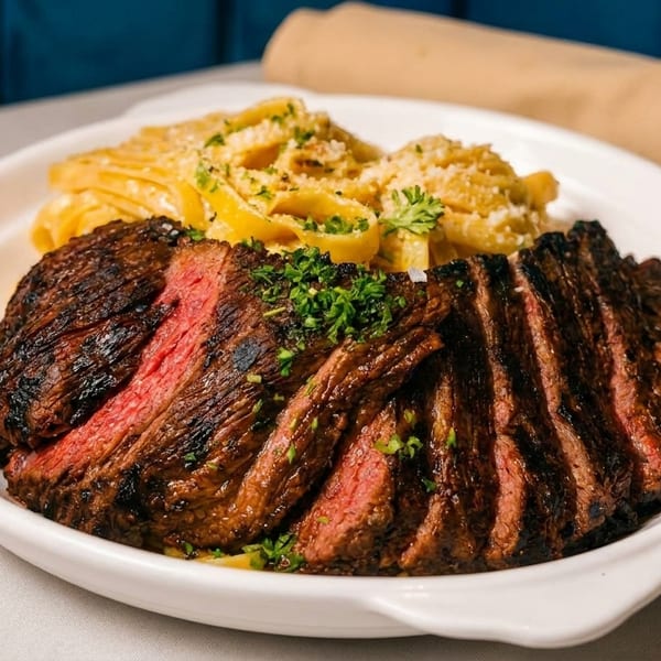 Sliced steak with a side of fettuccine pasta, garnished with parsley and parmesan cheese.