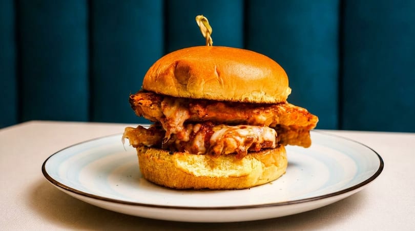 A cheesy chicken sandwich on a brioche bun, served on a white plate with blue accents.