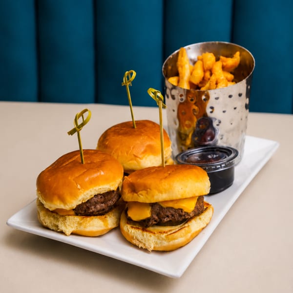 Three cheeseburger sliders with toothpicks, a side of fries in a metal cup, and dipping sauce.