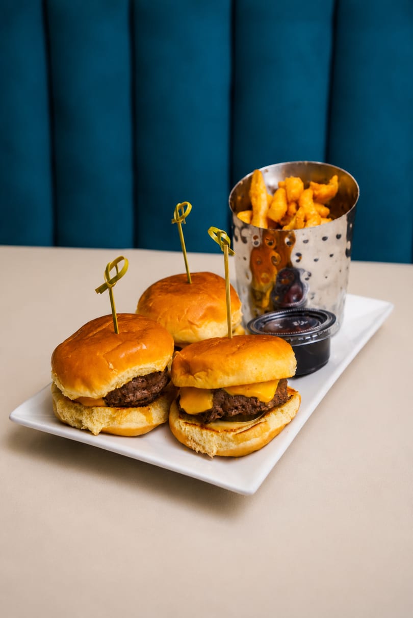 Three cheeseburger sliders with toothpicks, a side of fries in a metal cup, and dipping sauce.