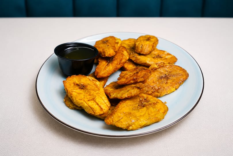 A light blue plate filled with golden-brown fried plantains and a small black cup of dipping sauce.