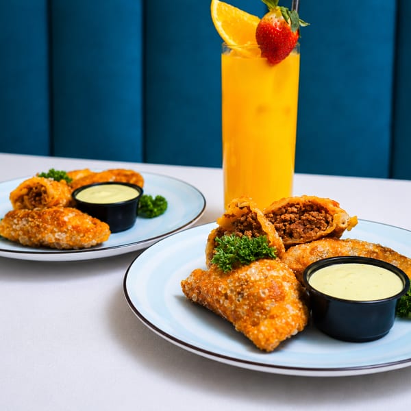 Two plates of fried empanadas with dipping sauce and a tall glass of orange juice.