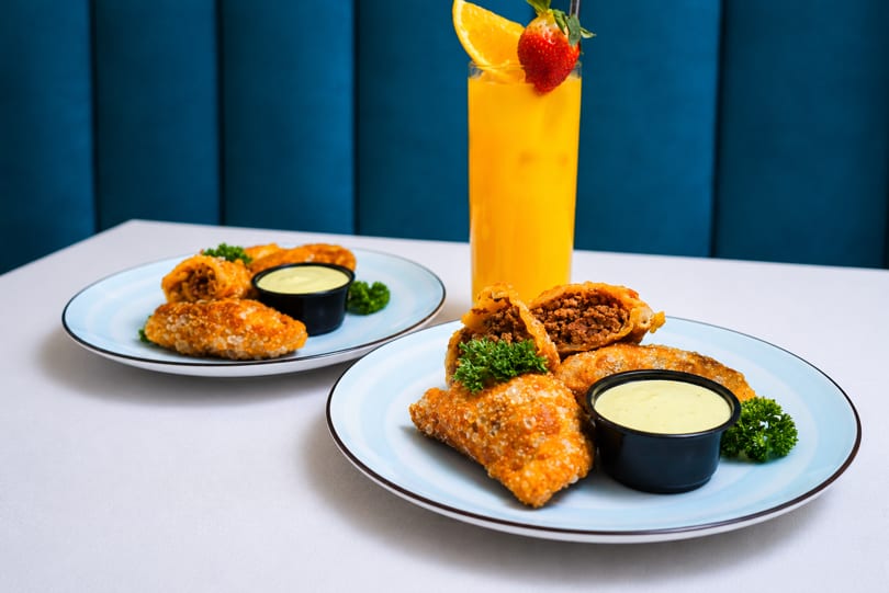 Two plates of fried empanadas with dipping sauce and a tall glass of orange juice.