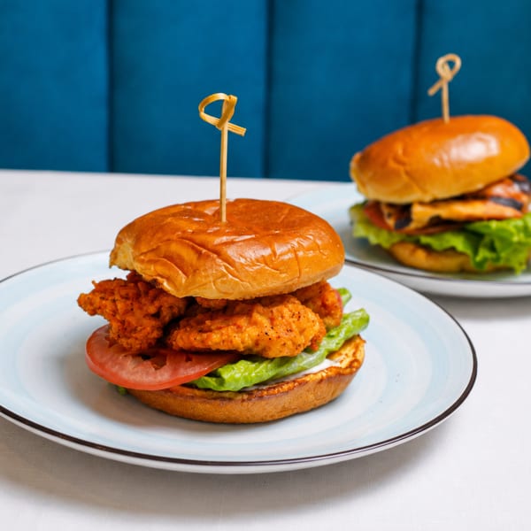 Two burgers on plates, one crispy chicken and one grilled chicken, on a white table with blue background.