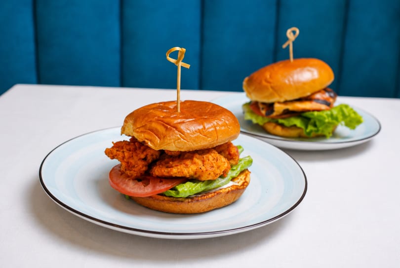Two burgers on plates, one crispy chicken and one grilled chicken, on a white table with blue background.